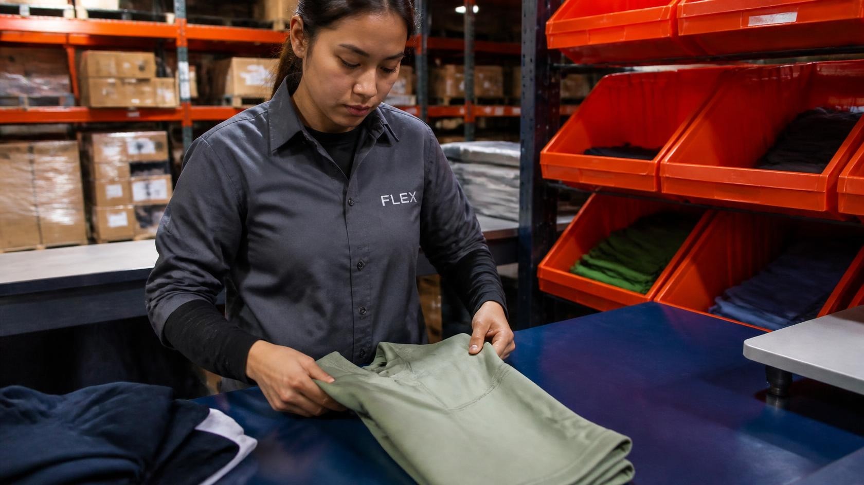 Garment packing associate wearing a slate gray shirt folding performance sportswear on a navy blue workbench with industrial orange storage bins.
